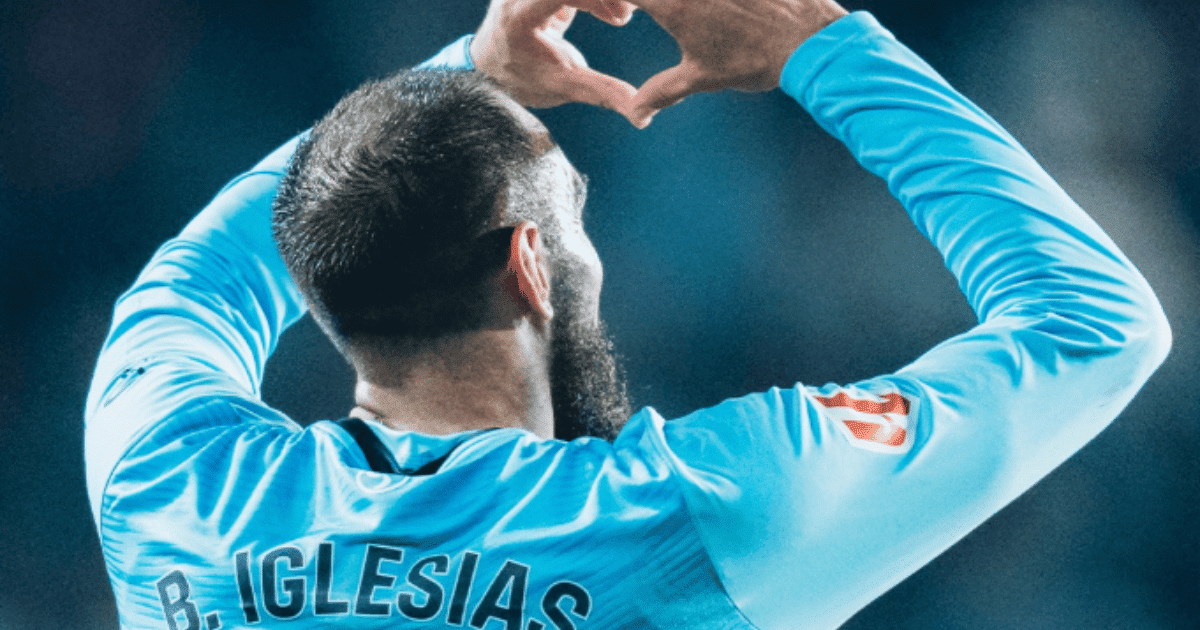 Primer plano del futbolista Borja Iglesias haciendo el signo del corazón con las manos frente a la grada.
