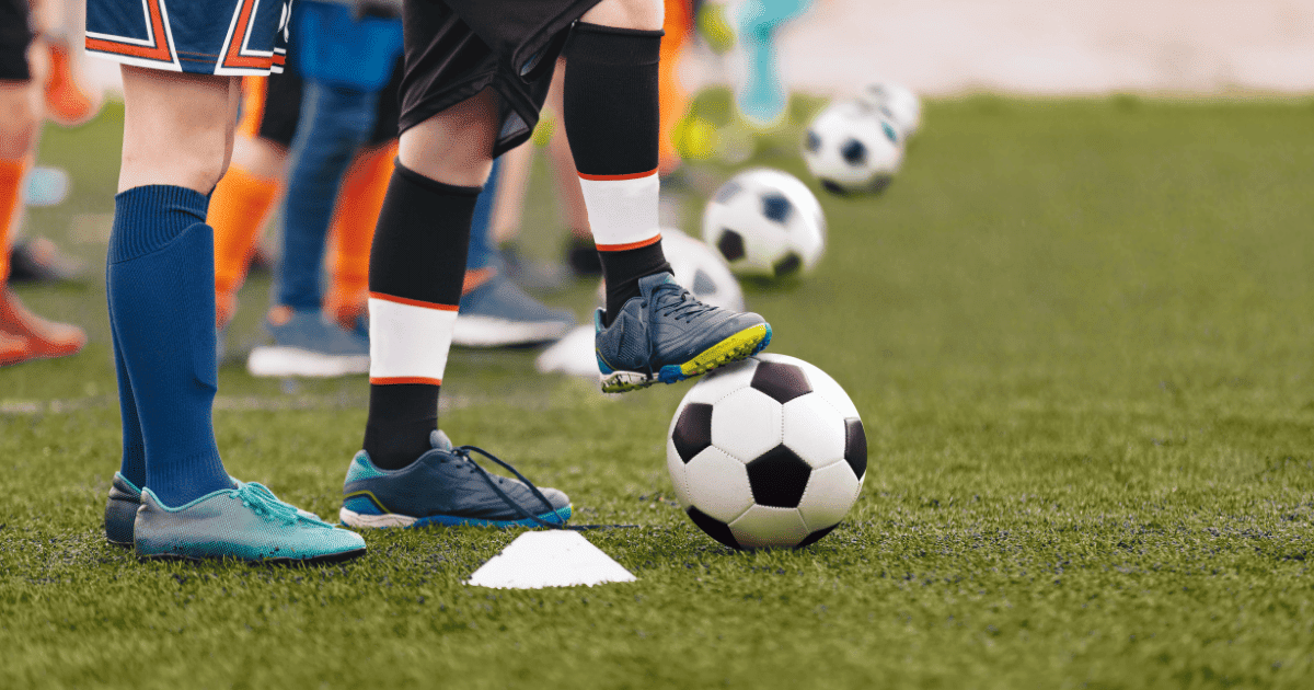Primer plano de pies de niños futbolistas con botas y balones sobre césped artificial en un entrenamiento.