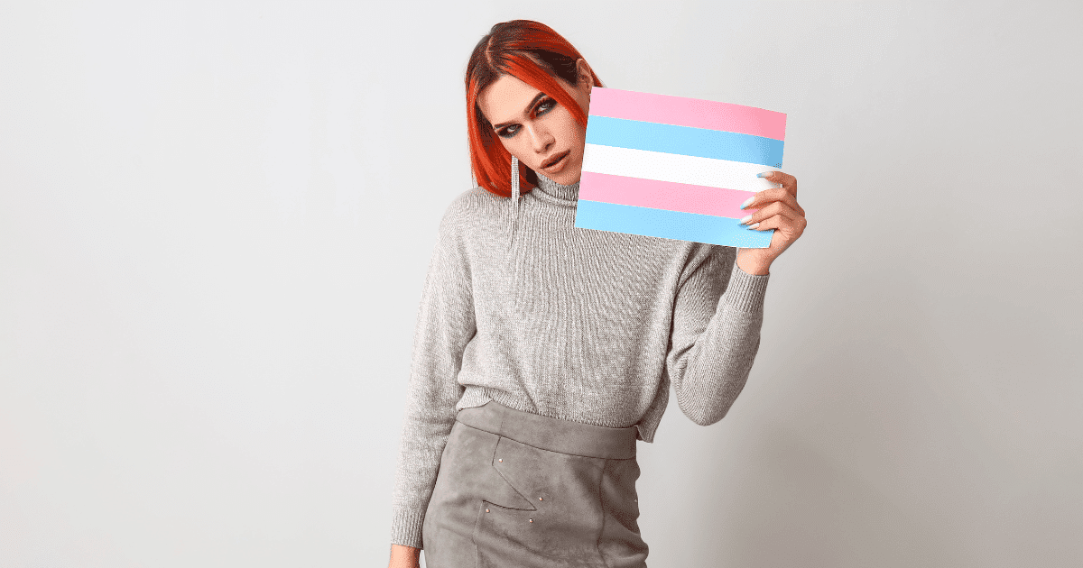Persona con cabello naranja brillante y maquillaje colorido sostiene con orgullo una bandera trans (franjas azul claro, rosa y blanco) frente a un fondo gris neutro. Imagen principal sobre la visibilidad trans en el empleo y las leyes de 2026.