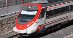 Fotografía en ángulo picado de un tren Renfe Cercanías de color blanco y rojo circulando por las vías cerca de una pared de piedra. Esta imagen ilustra la noticia sobre la denuncia de agresión tránsfoba ocurrida en la línea de Málaga a Torremolinos.