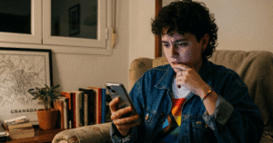 Persona joven sentada en un sofá mirando con preocupación su teléfono móvil en un entorno doméstico; la imagen representa la vulnerabilidad digital del colectivo LGTBIQ+ mencionada en el informe de GLAAD.