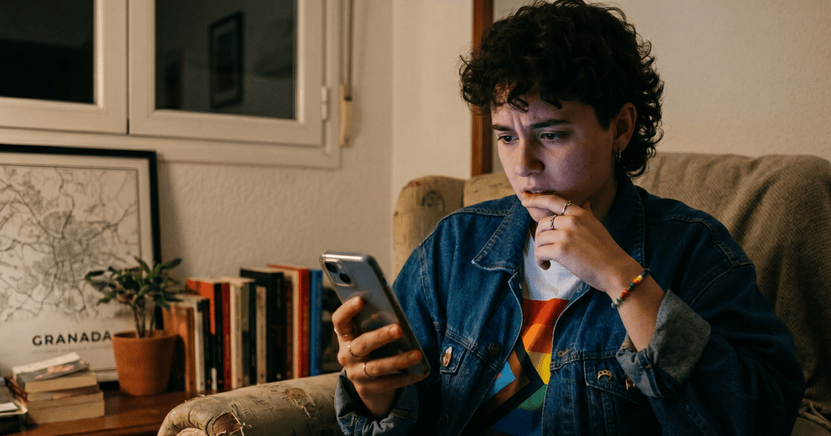 Persona joven sentada en un sofá mirando con preocupación su teléfono móvil en un entorno doméstico; la imagen representa la vulnerabilidad digital del colectivo LGTBIQ+ mencionada en el informe de GLAAD.