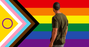 Plano posterior de una persona con camiseta verde contemplando la bandera del Orgullo Progresista Intersex inclusiva, que muestra franjas horizontales con los colores del arcoíris y un triángulo lateral con los colores trans, trazo intersex y bandas representativas de la diversidad.