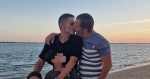 Dos hombres besándose en la playa mientras abrazan a sus dos hijos menores, con rostros pixelados para proteger su intimidad.