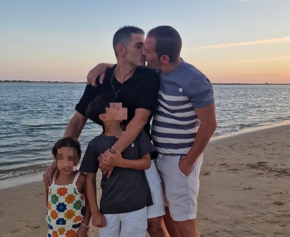 Fotografía en una playa donde una pareja de dos hombres se besa acompañados de sus dos hijos menores, cuya cara aparece pixelada para proteger su privacidad.