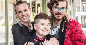 Tres personas sonrientes con diversas expresiones de género, maquillaje, pendientes y vestimenta que combina elementos femeninos y masculinos.